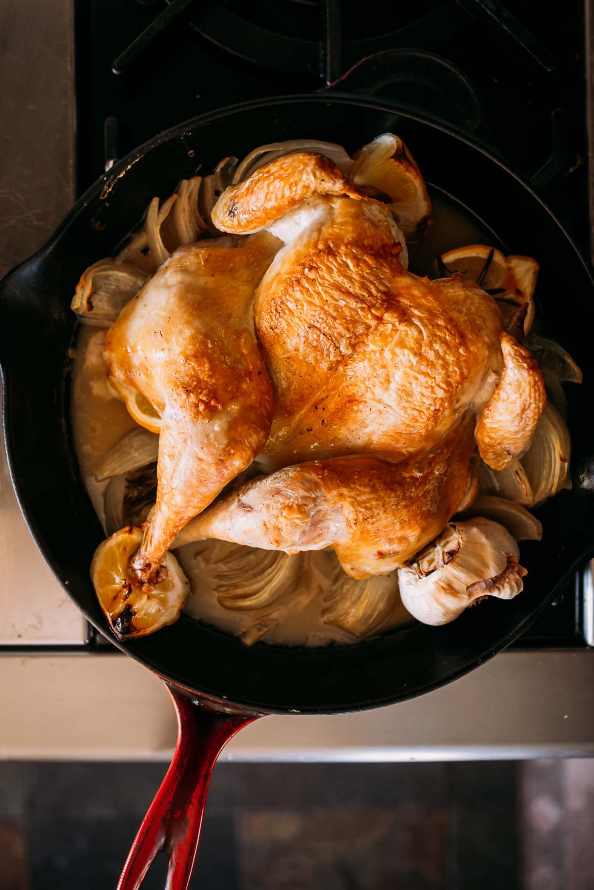 Roasted chicken with a golden-brown skin lies in a skillet surrounded by onions and garlic cloves.