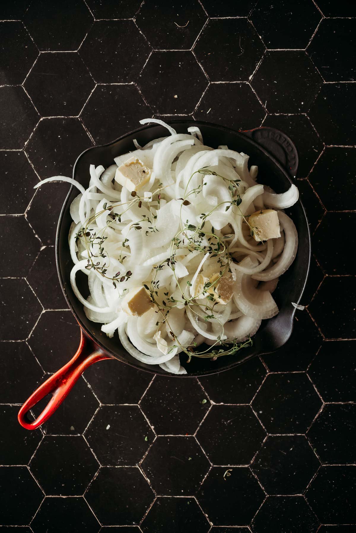 A pan with sliced raw onions, cubes of butter, and sprigs of thyme on a dark hexagonal-tiled surface.