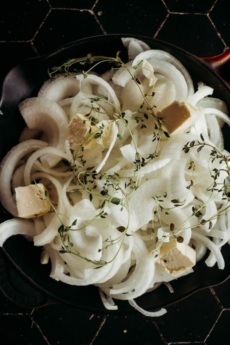 Sliced sweet onions with thyme sprigs and butter cubes in a skillet on a hexagonal tiled surface.