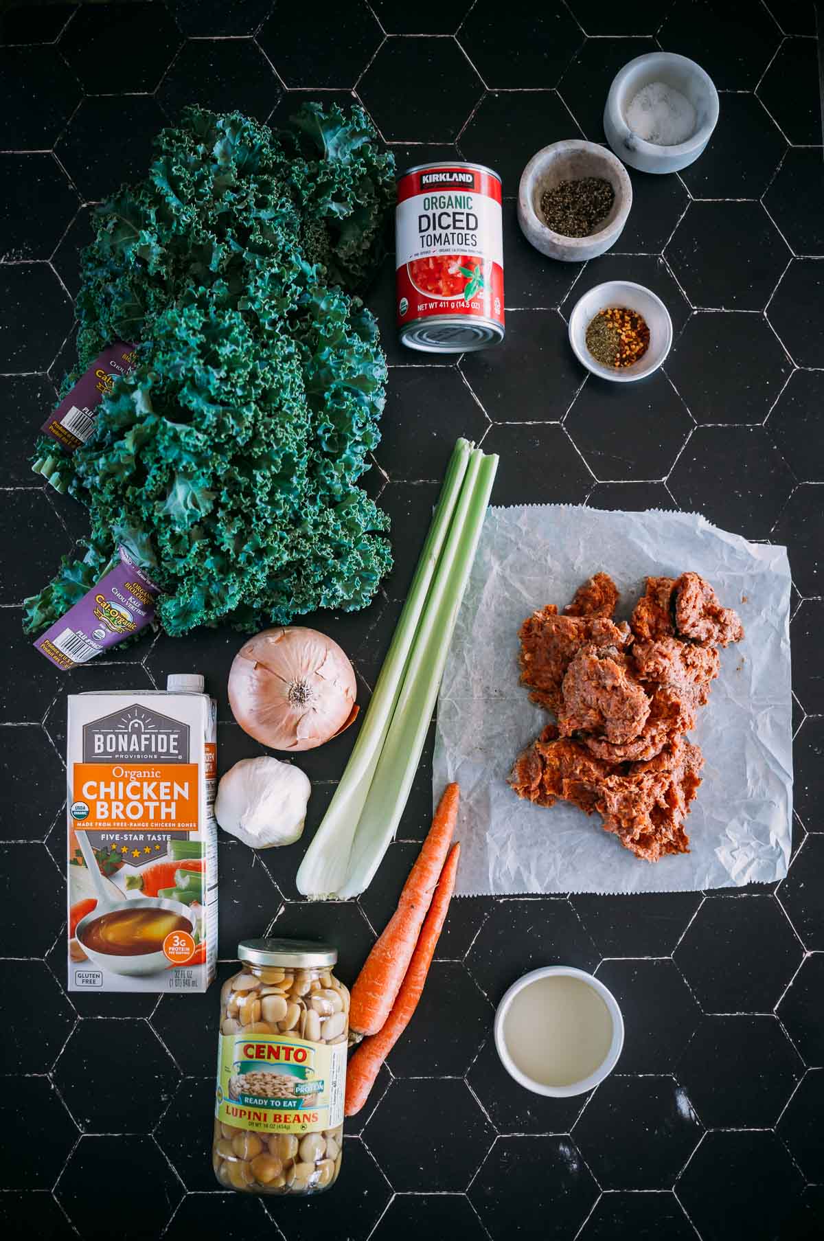 Ingredients on a black hexagonal tile surface: kale, canned tomatoes, spices, onion, chicken broth, celery, carrots, ground meat, beans, and white wine in a small bowl.