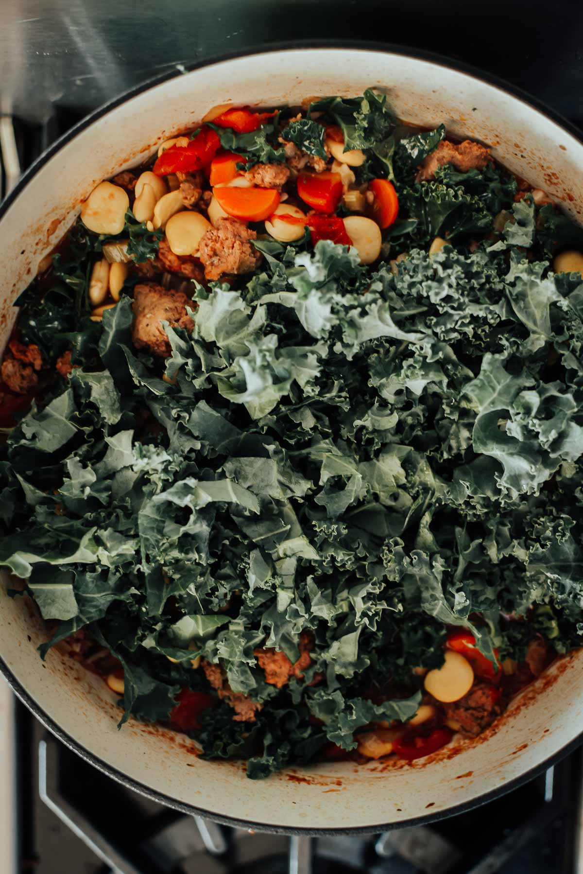 A pot on the stove contains ingredients like chopped kale, sliced carrots, red bell peppers, and beans in a soup.