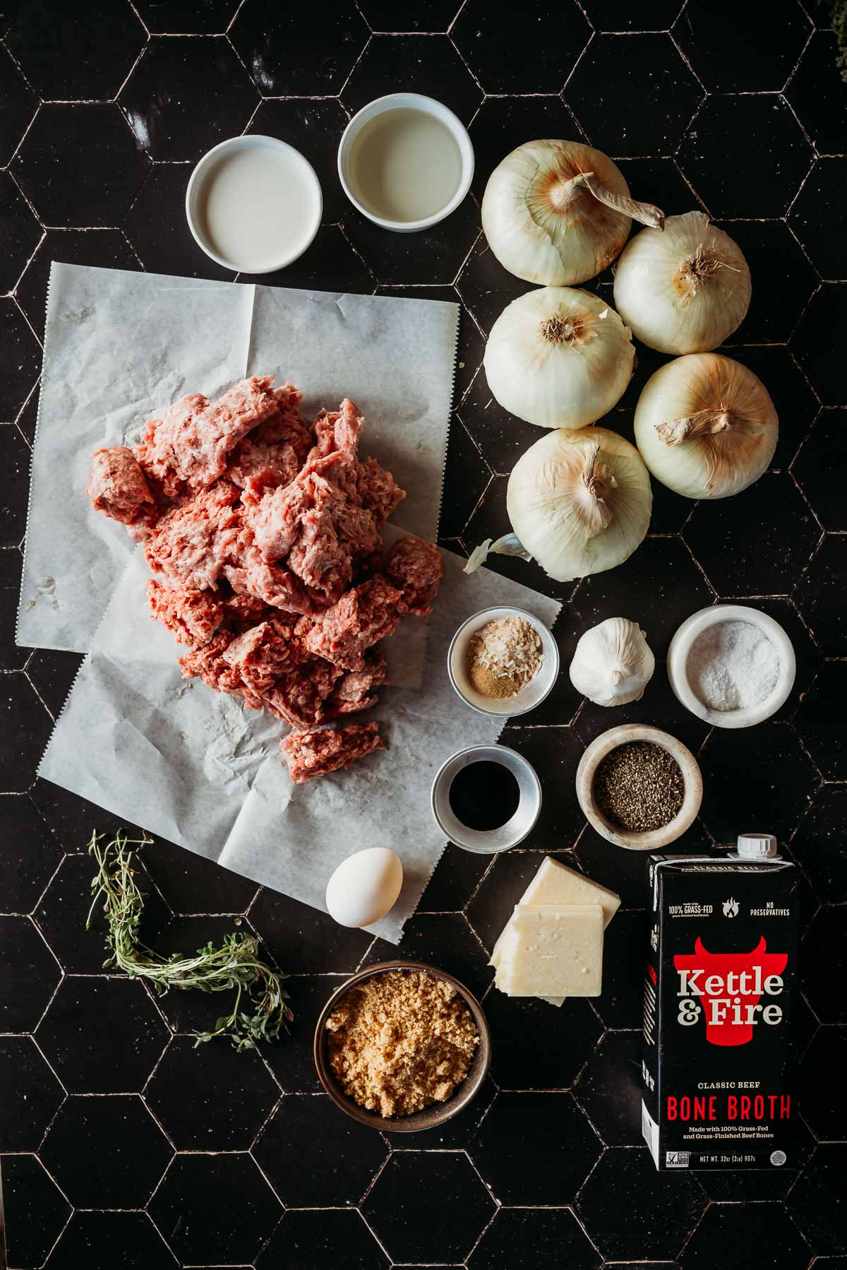 A black tile surface with ground meat, onions, egg, bone broth, herbs, spices, bouillon, and seasonings arranged around.