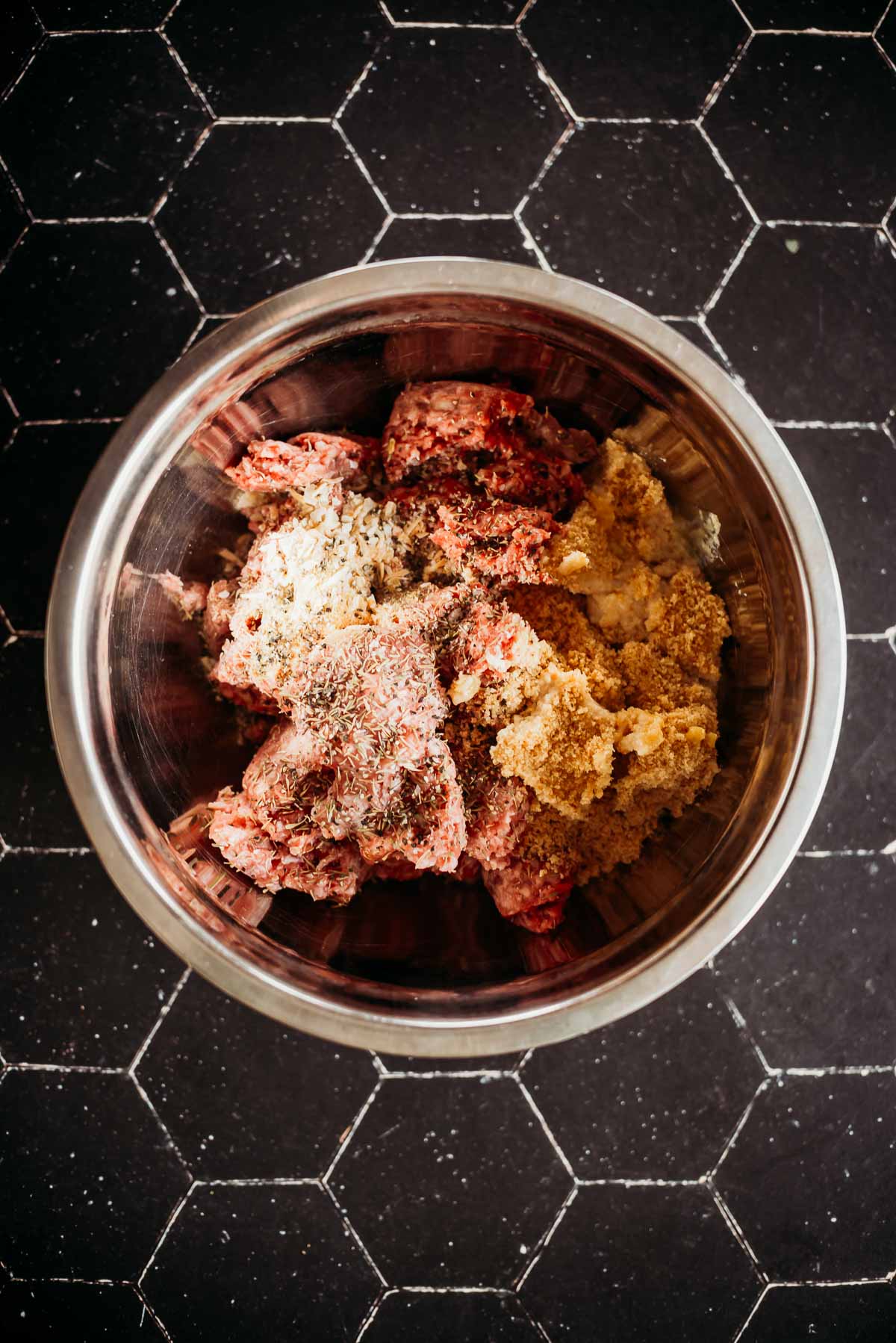 Bowl with ground meat mixed with spices and breadcrumbs on a tiled surface.