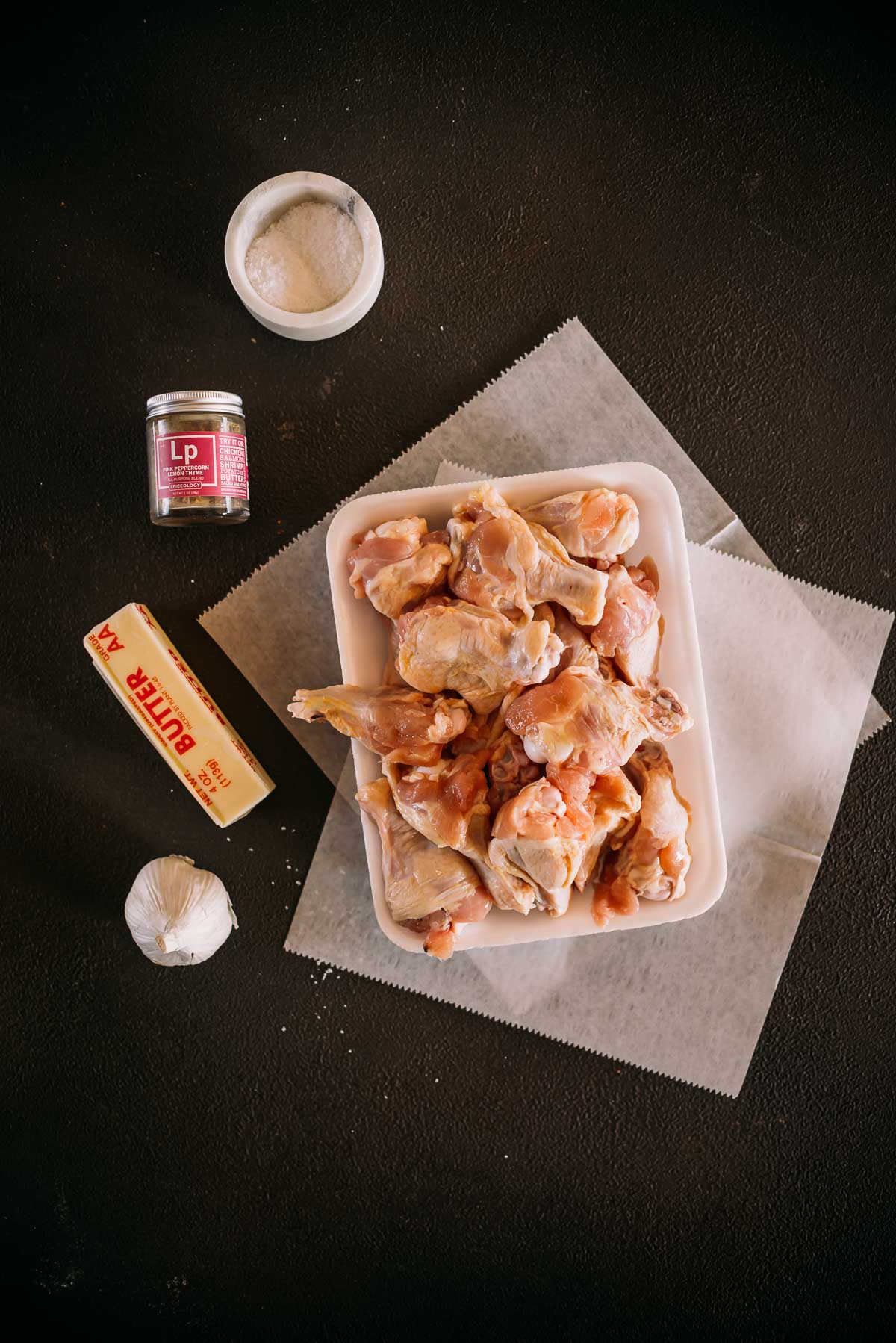 A tray of raw chicken wings is placed on parchment paper. Surrounding it are a stick of butter, a bulb of garlic, a jar of seasoning, and a container of salt on a dark surface.