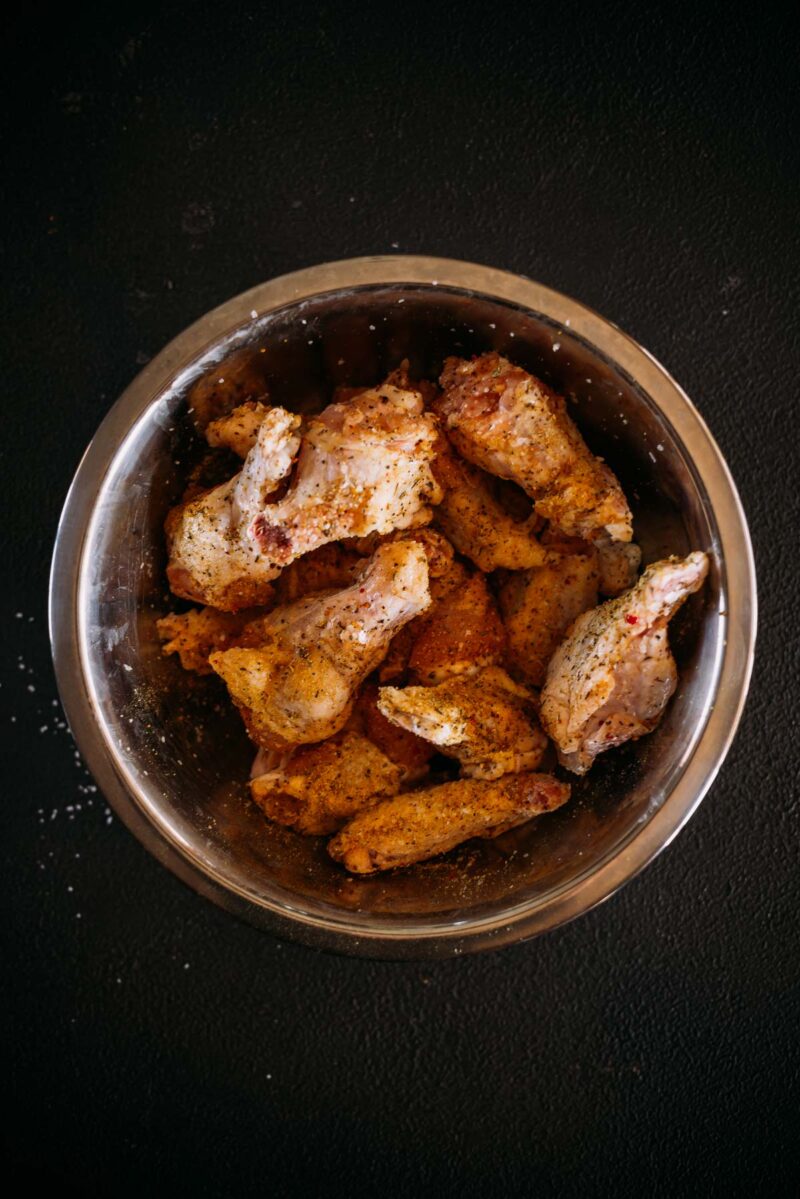 A metal bowl filled with seasoned, cooked chicken wings on a dark surface.