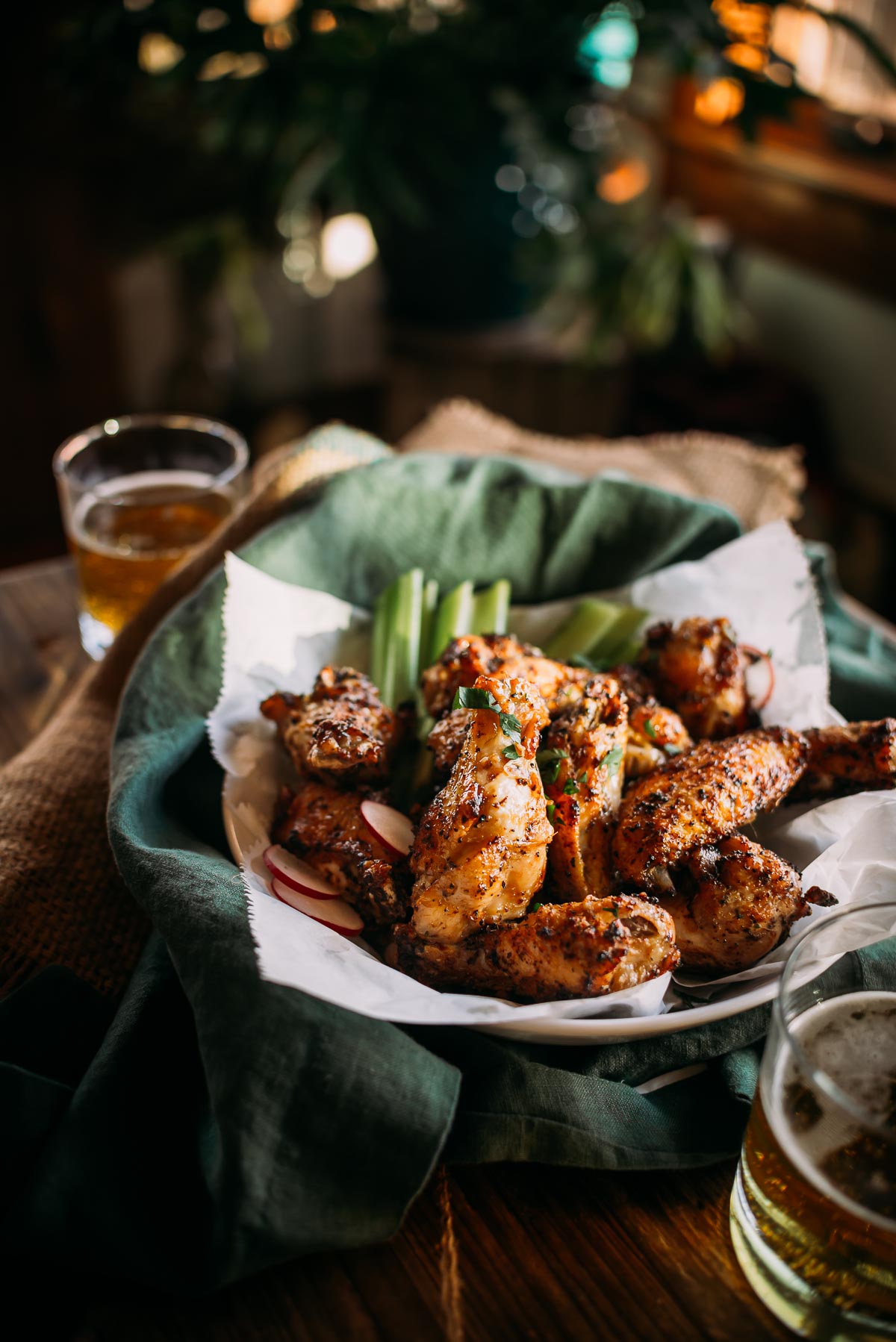 A plate of lemon pepper chicken wings with herbs, garnished with celery sticks and radish slices, placed on a green cloth beside two glasses of beer.