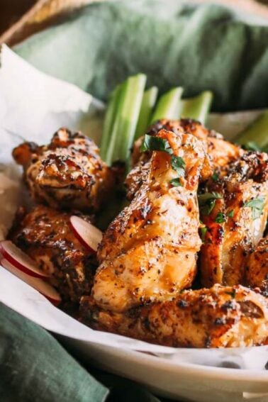 Plate of seasoned and grilled chicken wings with celery sticks, set on a green cloth. A glass of amber-colored liquid is in the background.