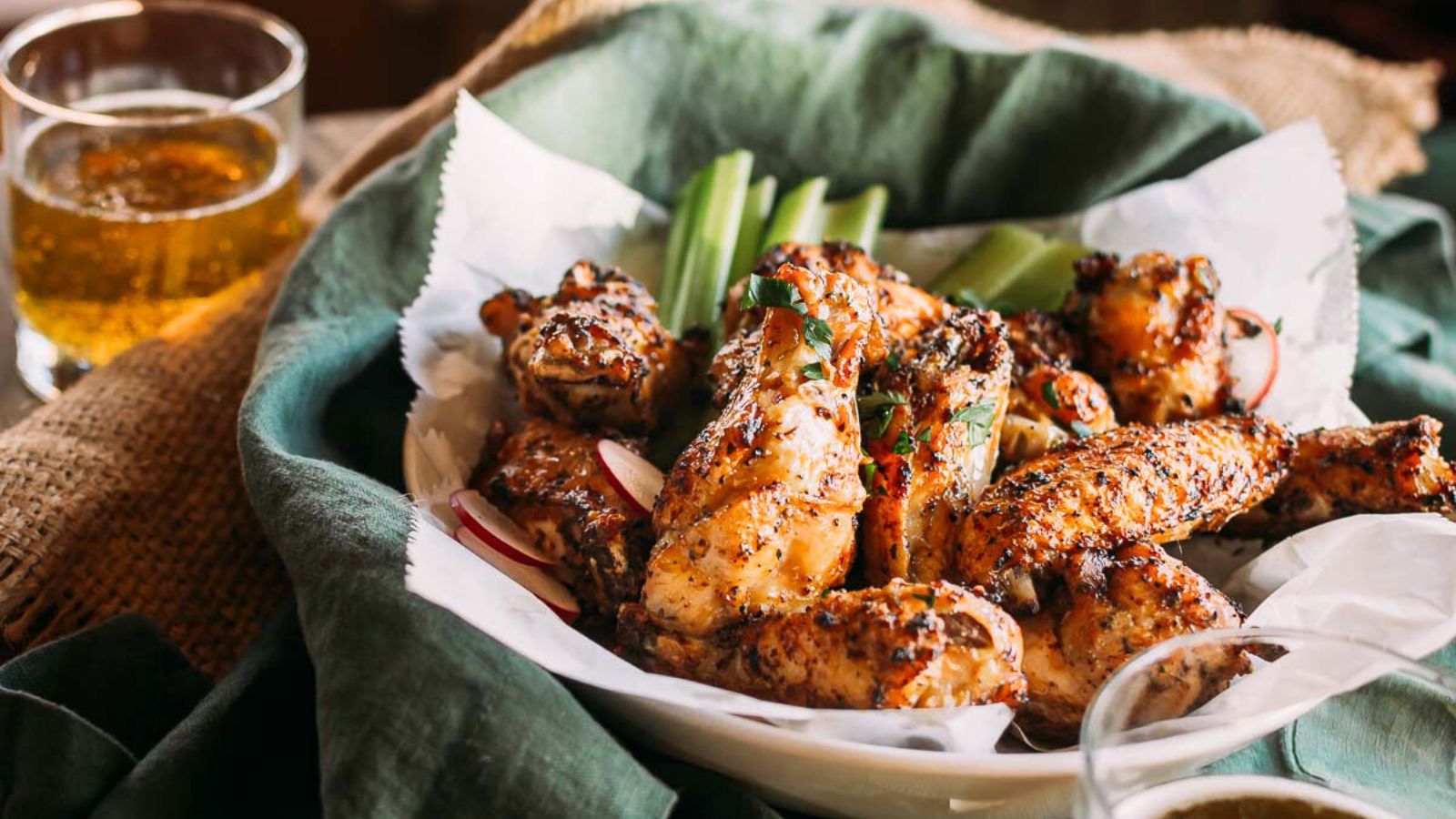 Plate of seasoned and grilled chicken wings with celery sticks, set on a green cloth. A glass of amber-colored liquid is in the background.