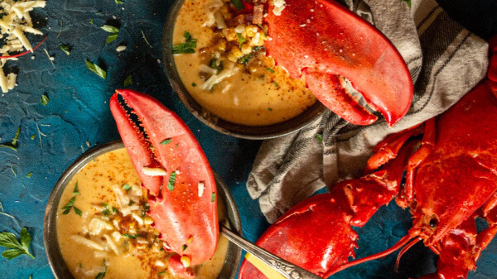 Two bowls of creamy soup topped with lobster claws sit elegantly beside a whole cooked lobster on a blue surface, perfect for surf and turf recipes for Valentine's Day.