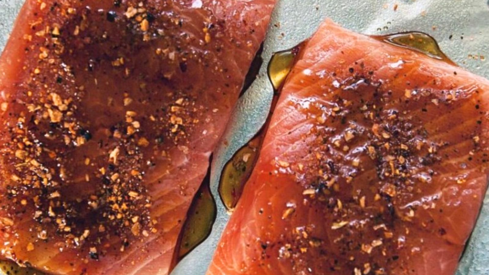 Two raw salmon fillets, perfect for Valentine's Day, are seasoned with spices and herbs, drizzled with oil, and displayed on a light-colored surface. Ideal for surf and turf recipes, these fillets promise a blend of flavors to elevate your special meal.