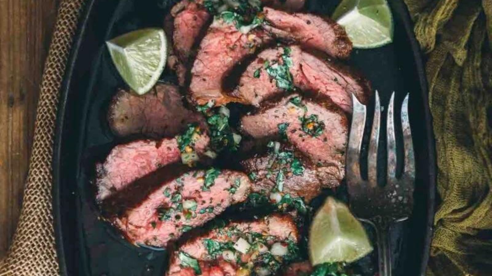 Grilled steak slices on a black platter, complemented by the essence of surf and turf recipes for Valentine's Day, are garnished with chopped herbs, onions, and lime wedges, with a fork on the side.