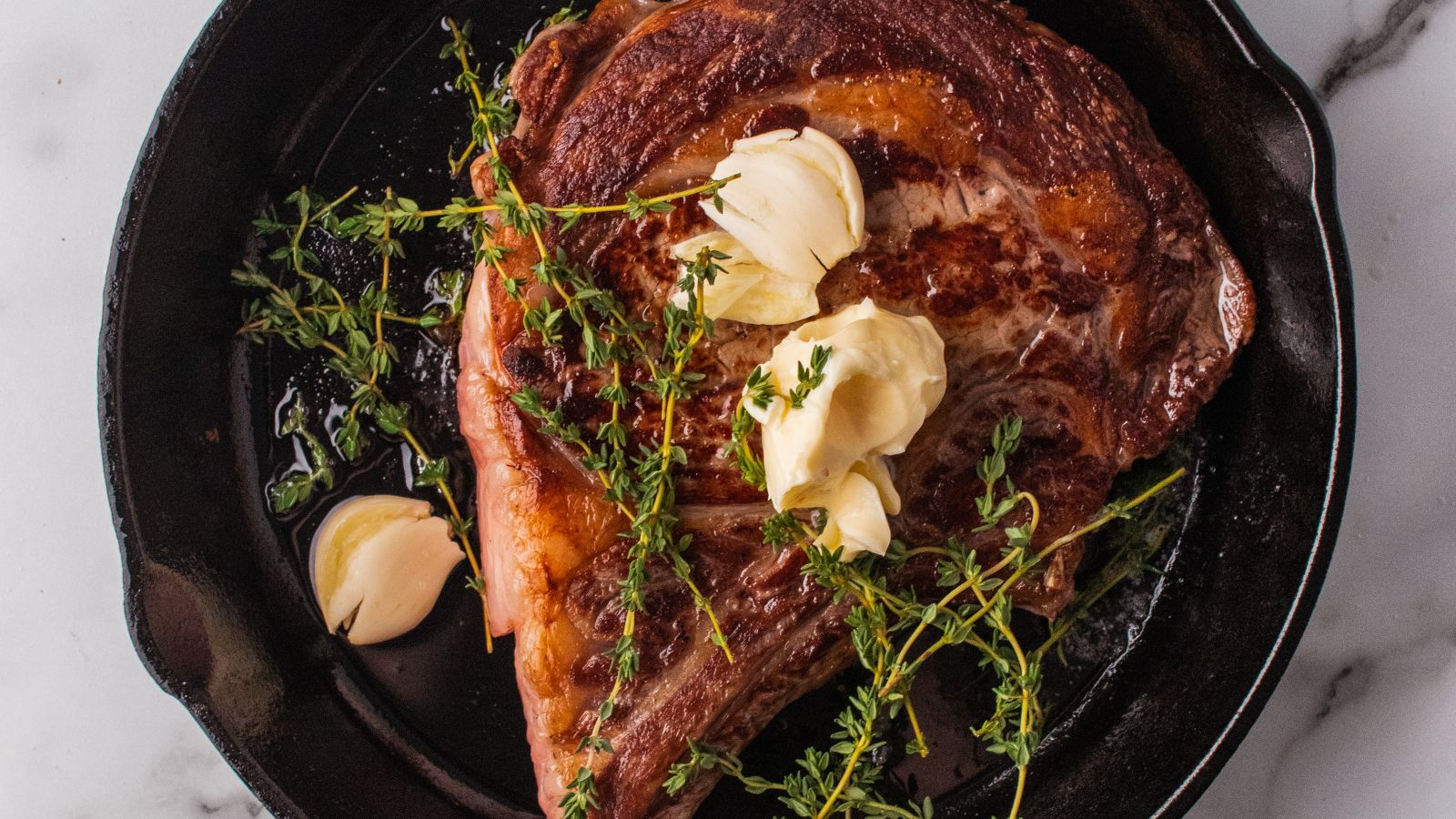 A cooked steak in a cast iron pan, topped with herbs, garlic, and butter, makes for an elegant surf and turf recipe perfect for Valentine's Day.