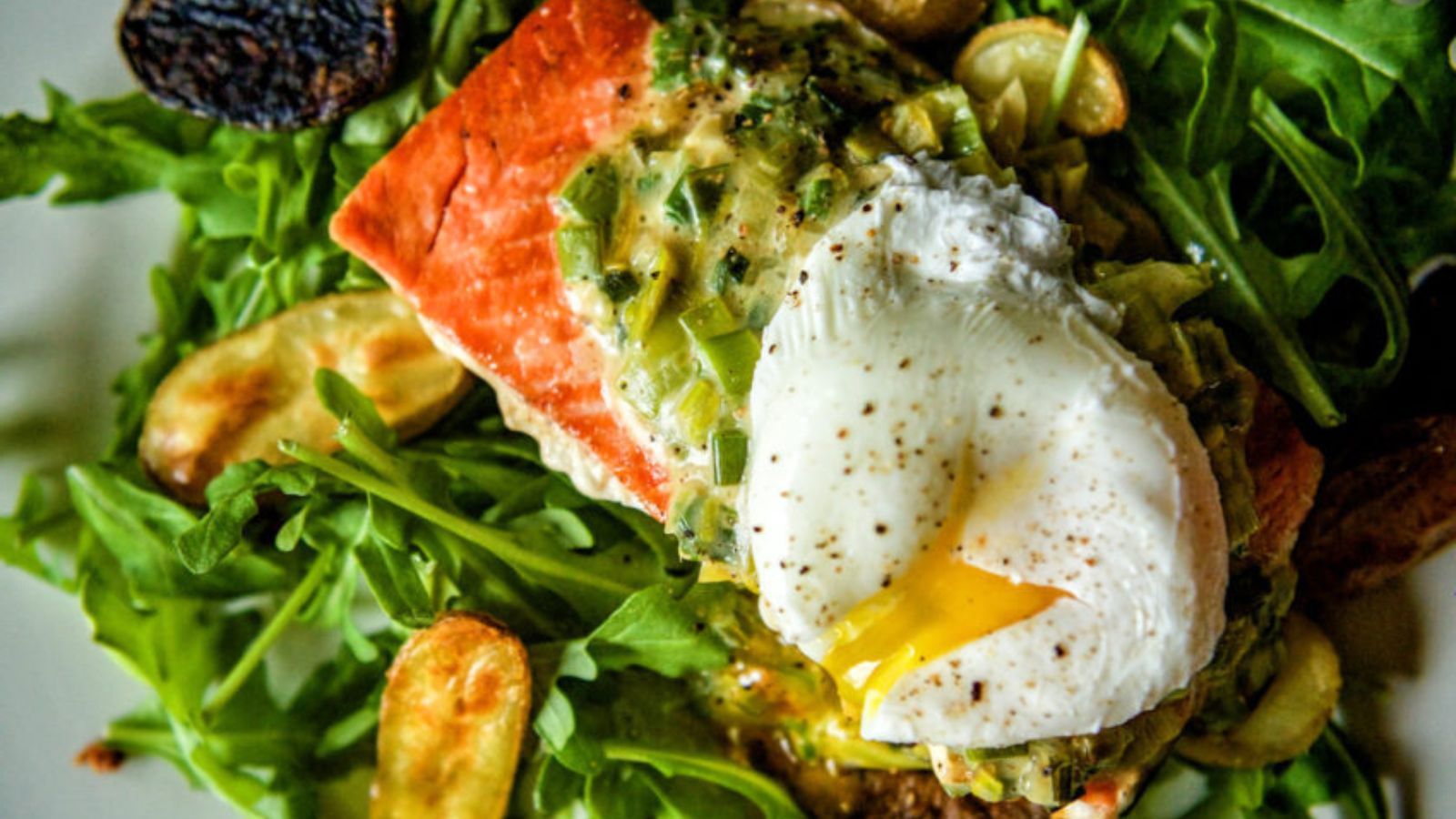 A poached egg with a runny yolk crowns grilled salmon, all elegantly arranged on a bed of arugula and herbs, complemented by roasted potatoes—an exquisite surf and turf recipe perfect for Valentine’s Day.