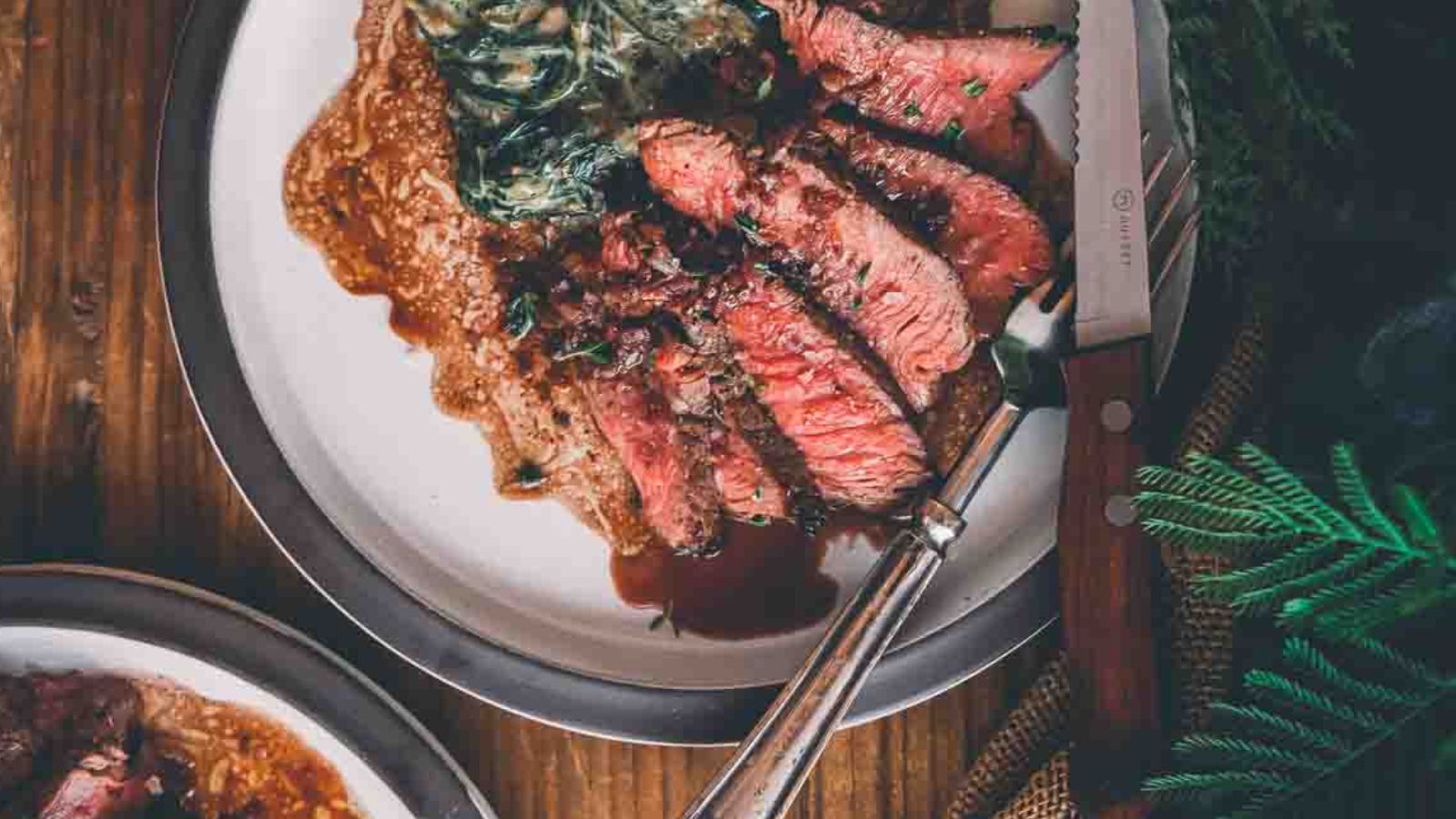 Sliced medium-rare steak on a plate with greens, sauce, and a steak knife on a wooden table.