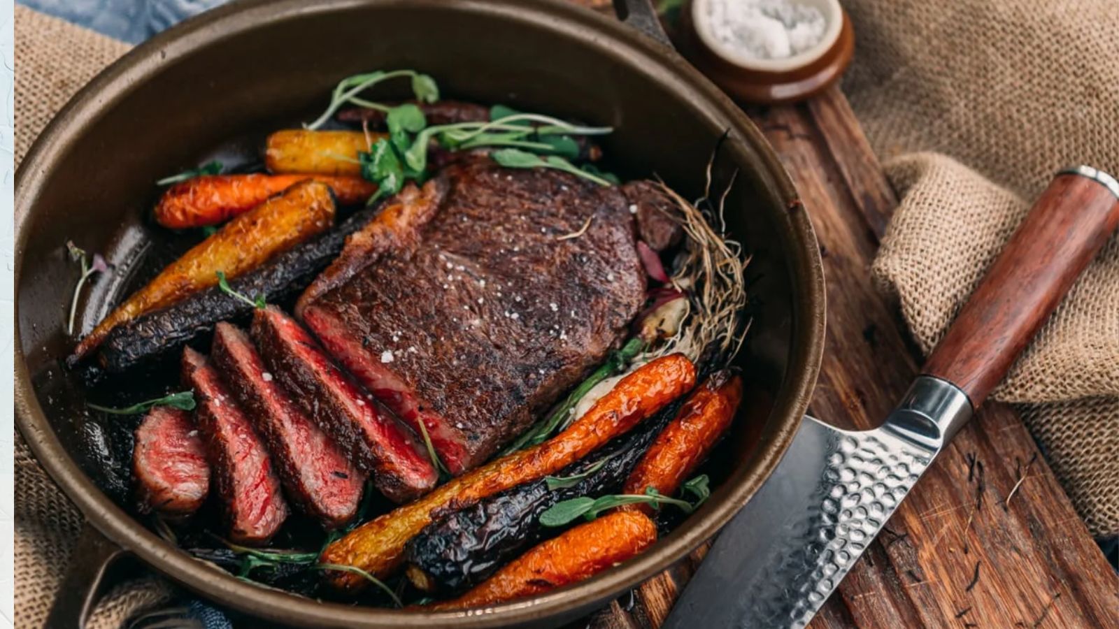 A cooked steak in a pan, partially sliced, pairs with roasted carrots and herbs—a perfect dish for surf and turf recipes for Valentine's Day. A knife rests nearby on the wooden surface.