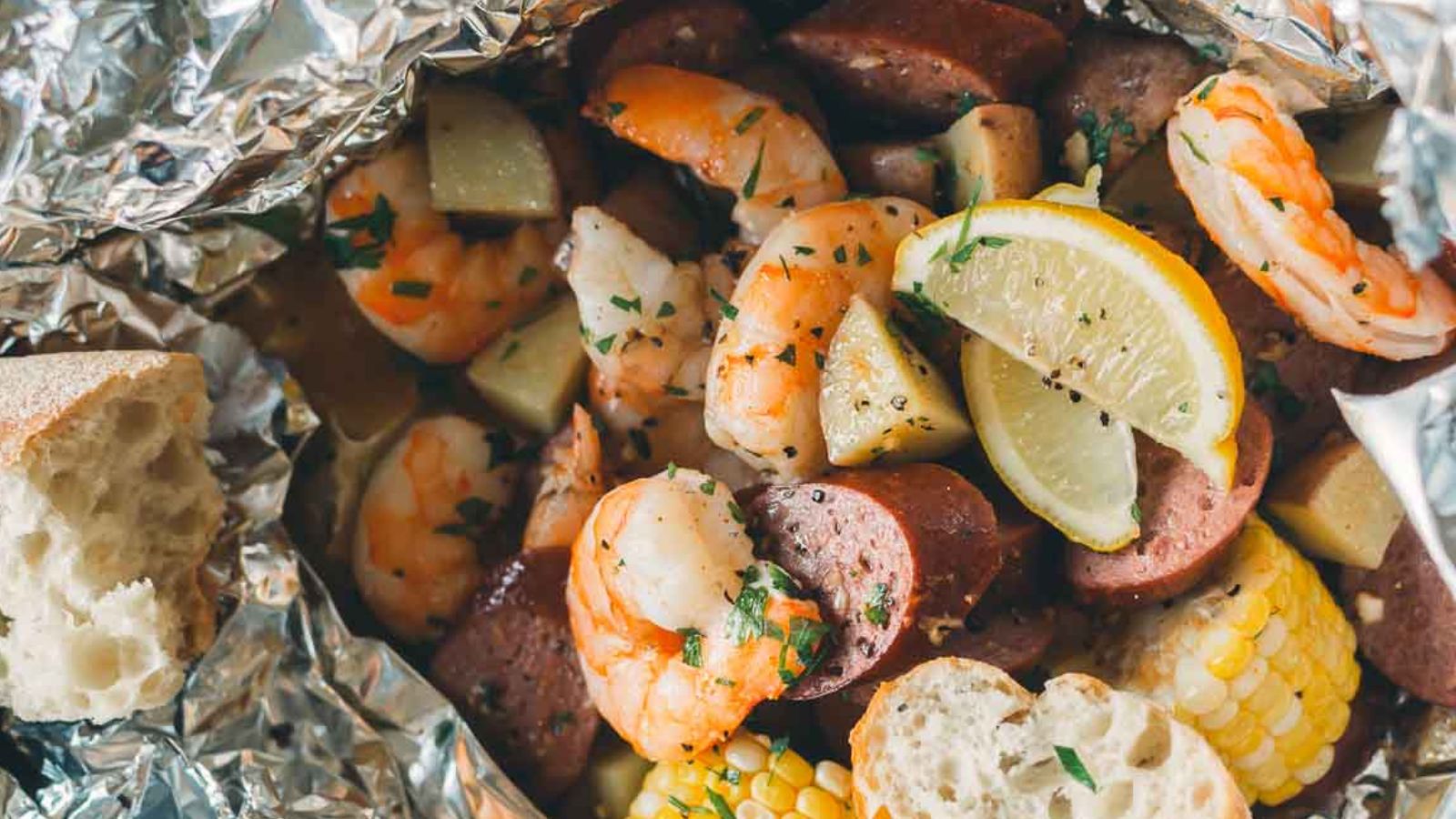 Foil packet containing shrimp, sausage, chopped potatoes, corn, and lemon wedges, garnished with herbs. Bread slices are visible on the side.