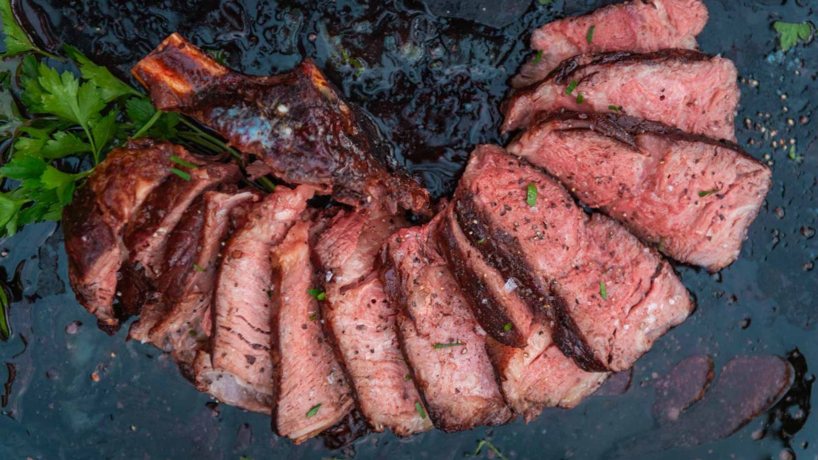 Sliced medium-rare steak, a perfect element for surf and turf recipes, is elegantly arranged on a dark surface with herbs on the side.