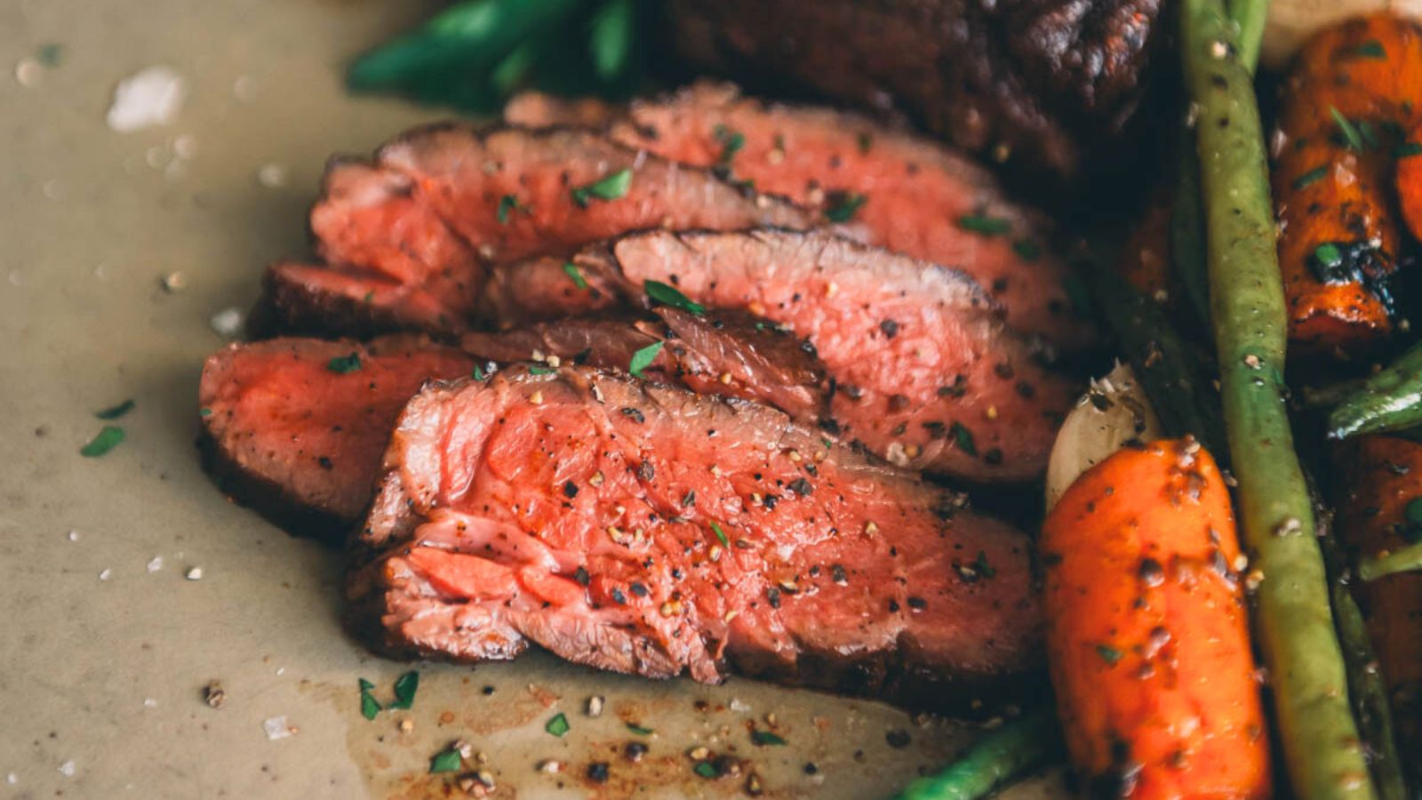 Slices of medium-rare steak garnished with herbs are artfully paired with roasted carrots and green beans on a plate, offering an elegant twist on classic surf and turf recipes for Valentine's Day.