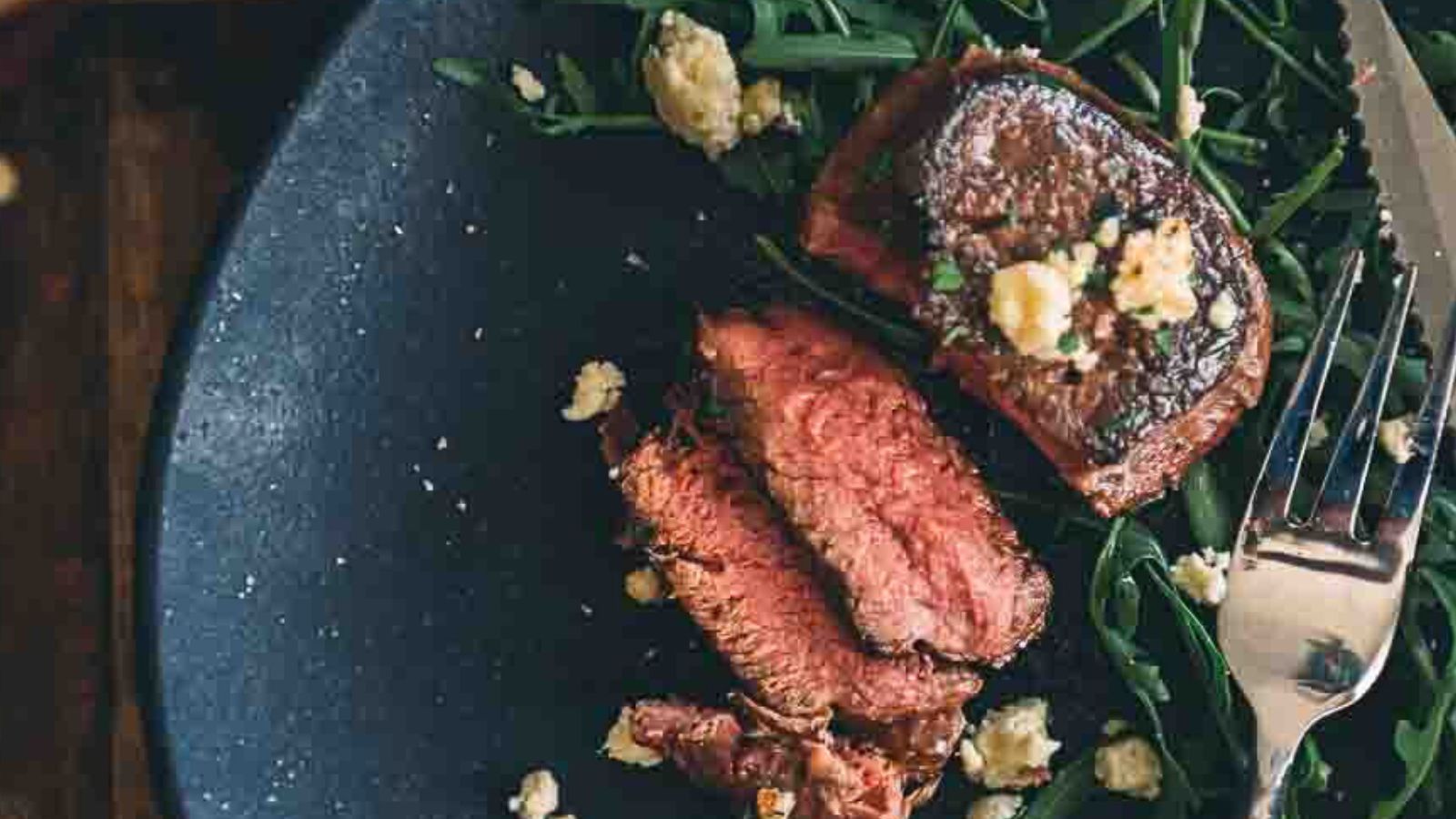 Sliced medium-rare steak on a bed of greens with crumbled cheese, served on a dark plate with a fork and knife on the side.