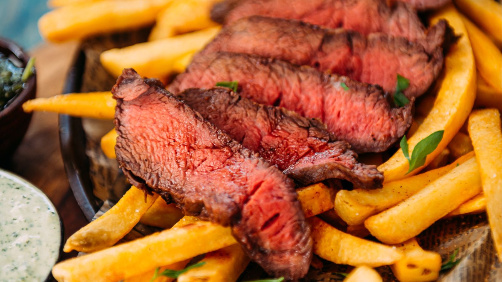 Slices of medium-rare steak on a bed of golden fries, garnished with small green herbs, crafts the perfect surf and turf recipe for Valentine's Day. Accompanied by a dish of green sauce on the side.