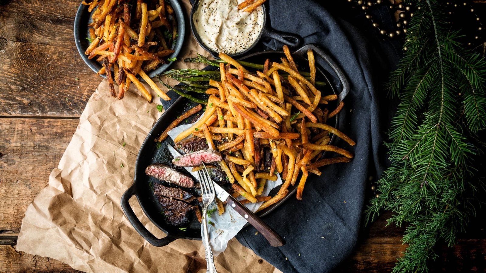 A plate of sliced steak with fries sits in a cast iron skillet, accompanied by a bowl of extra fries and a sauce cup, all set on a wooden table adorned with paper, cloth, and pine branches. Perfect for experimenting with surf and turf recipes for Valentine's Day.