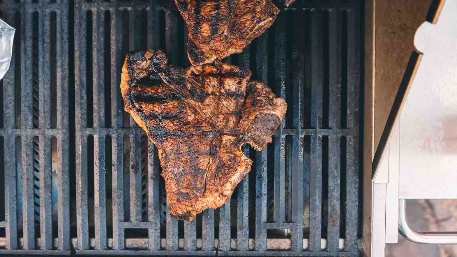 Two T-bone steaks with prominent grill marks sizzle on the grill, perfect for an indulgent surf and turf recipe ideal for Valentine's Day.