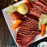 Plate of sliced corned beef, cabbage, carrots, potato, and onion on a white square plate. Sliced bread and a dish of sour cream are nearby. Text reads "Corned Beef & Cabbage.