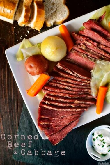 Plate of sliced corned beef, cabbage, carrots, potato, and onion on a white square plate. Sliced bread and a dish of sour cream are nearby. Text reads "Corned Beef & Cabbage.