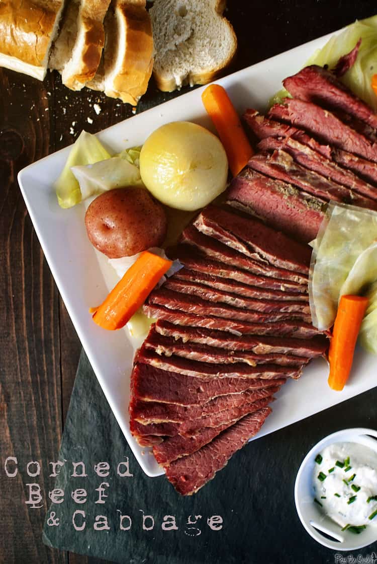 Plate of sliced corned beef, cabbage, carrots, potato, and onion on a white square plate. Sliced bread and a dish of sour cream are nearby. Text reads "Corned Beef & Cabbage.