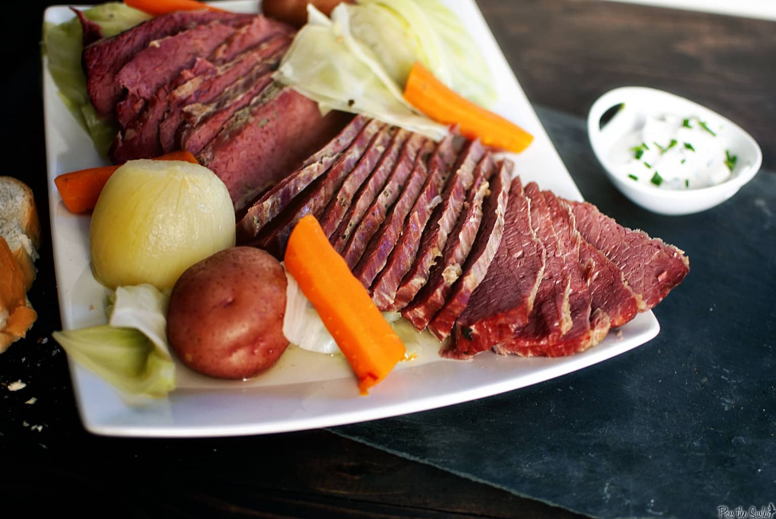 Sliced corned beef with carrots, potatoes, cabbage, and onions on a platter. A small dish of creamy sauce with chives is beside it.