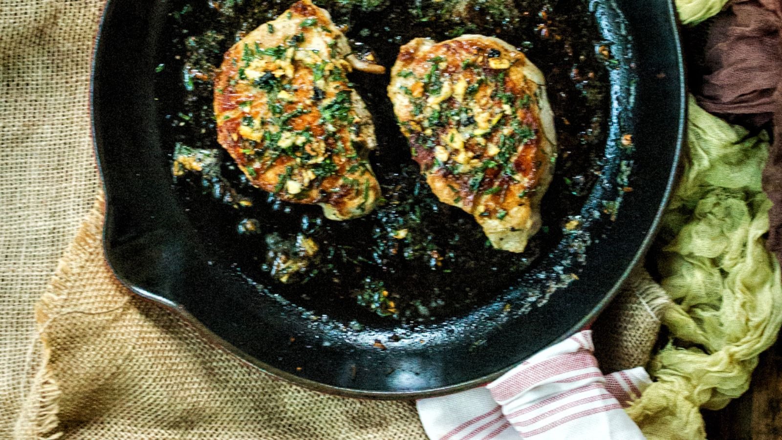 Two cooked chicken breasts seasoned with herbs in a black skillet on a textured cloth background.