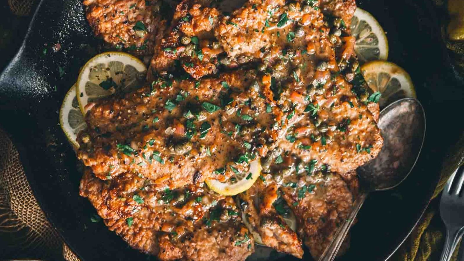 Cooked chicken with sauce, garnished with parsley and capers, served on a cast iron skillet with lemon slices and a metal spoon.