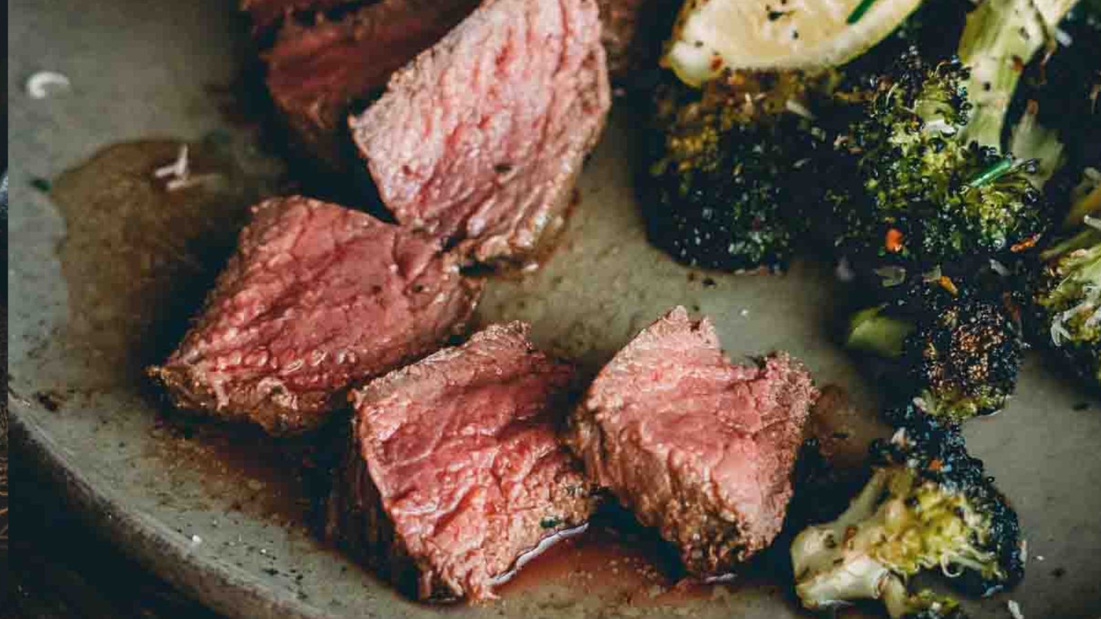 Slices of medium-rare steak on a plate with roasted broccoli and potato wedges.