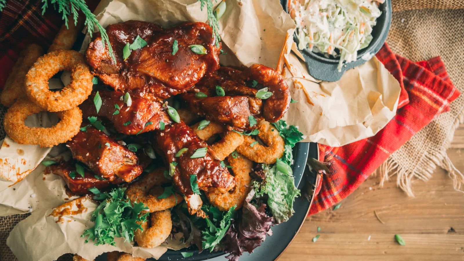 Plate of barbecue chicken wings with onion rings, garnished with greens, and a side of coleslaw on rustic burlap.