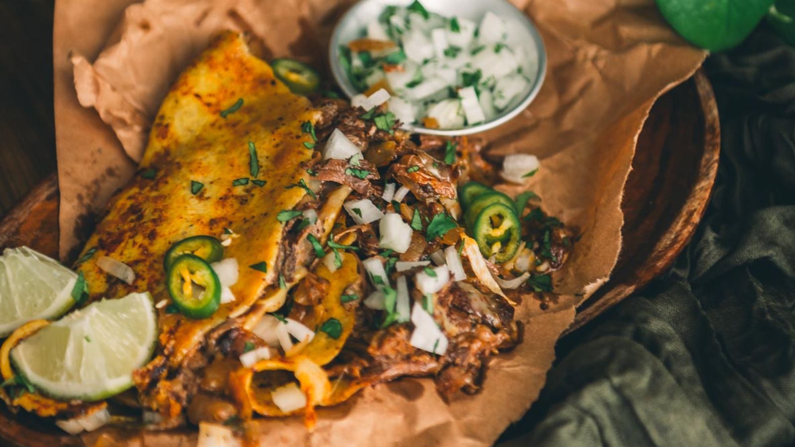 A birria taco filled with beef, onions, and cilantro, served with lime wedges and a small bowl of chopped onions and herbs on a wooden plate lined with parchment paper.