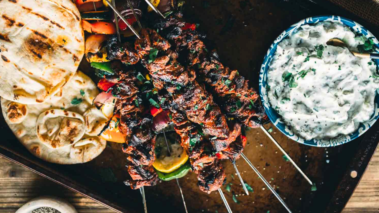 Grilled lamb and vegetable skewers on a tray, served with pita bread and a bowl of creamy herb dip.