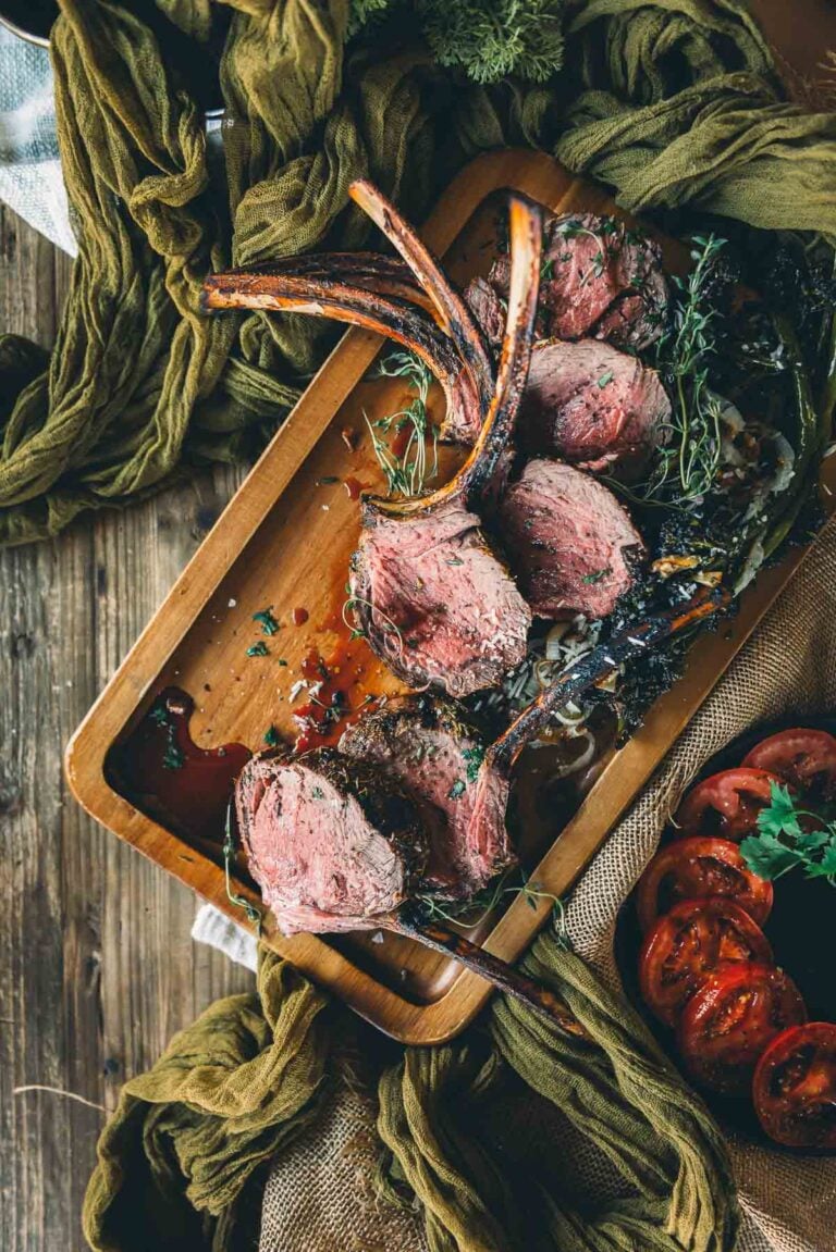 A wooden tray holds sliced grilled rack of elk garnished with herbs, set on a rustic table with green cloth and sliced tomatoes nearby.