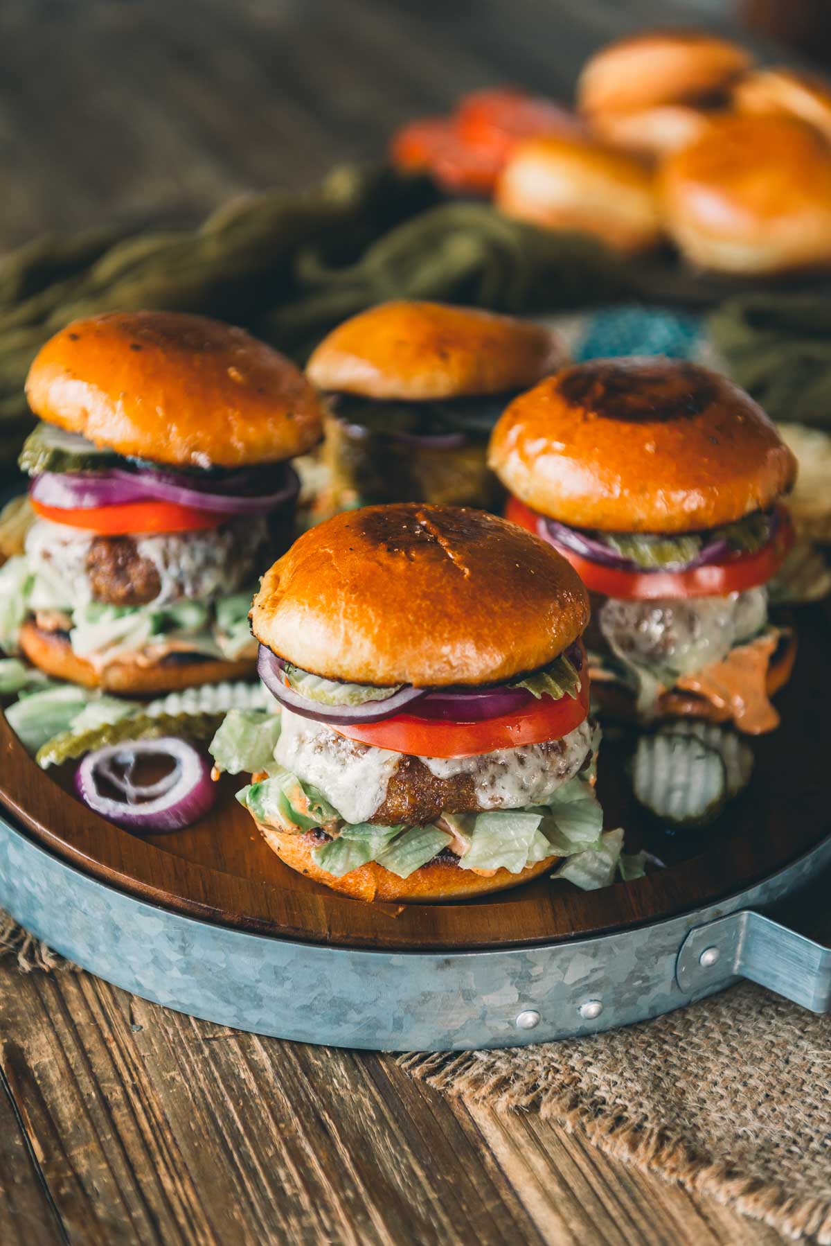 Four cheeseburgers with lettuce, tomato, pickles, and onions on brioche buns are arranged on a round wooden tray.