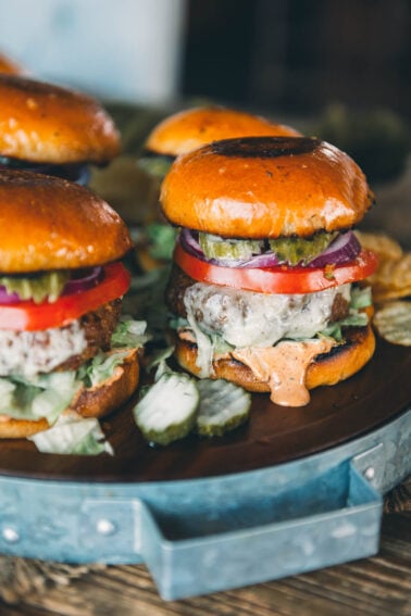 A turkey burger with lettuce, tomato, red onion, pickles, and sauce in a brioche bun, served on a round metal tray with sliced pickles and another burger in the background.