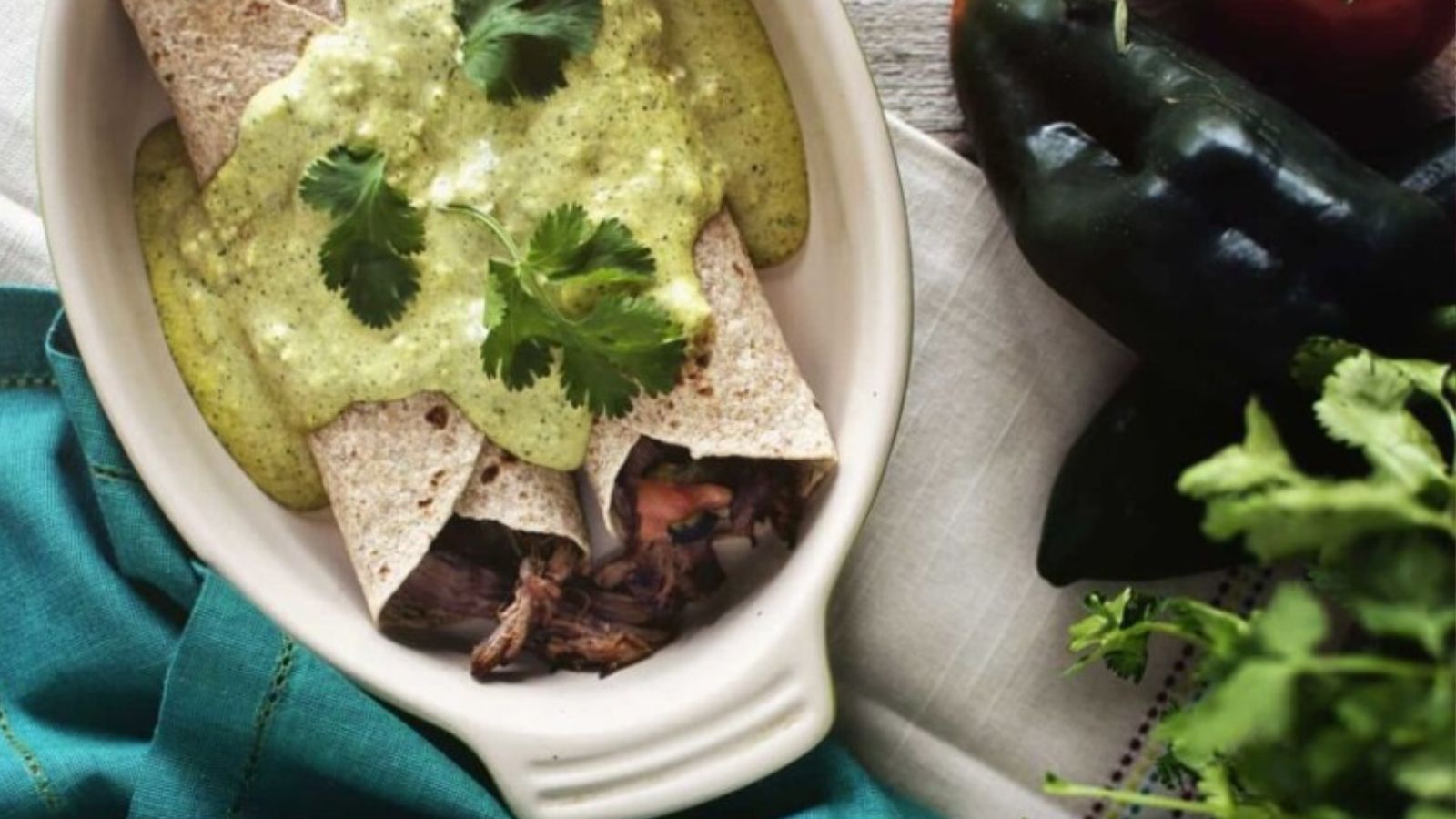 A dish with two rolled tortillas filled with meat, topped with green sauce and cilantro, next to fresh peppers and herbs on a blue cloth.