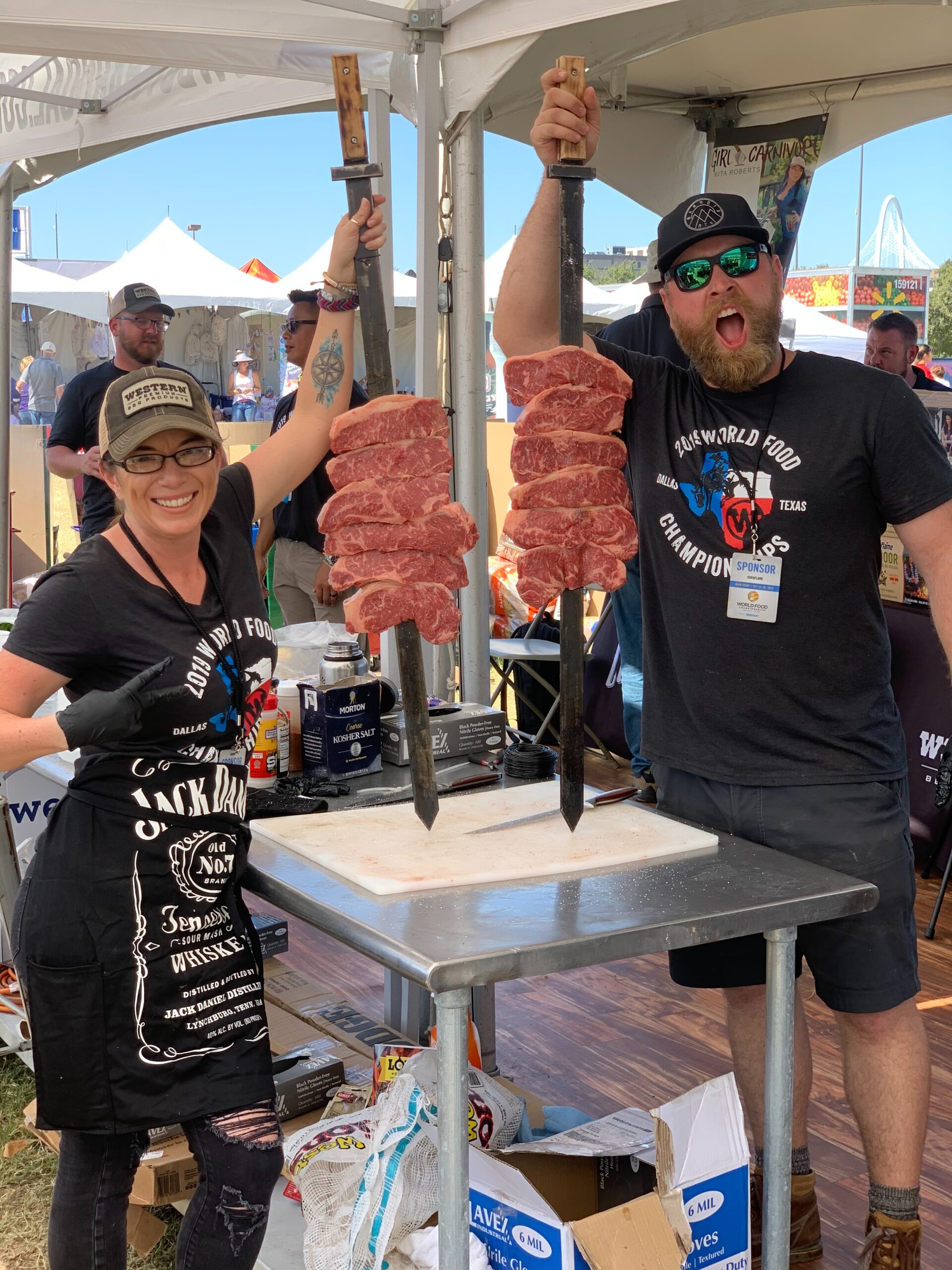 Two people at an outdoor event hold large skewers with several pieces of raw meat, standing behind a table under a tent.