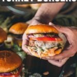 Hands holding an air fryer turkey burger with melted cheese, tomato, lettuce, and sauce; more burgers and pickles on a tray below.