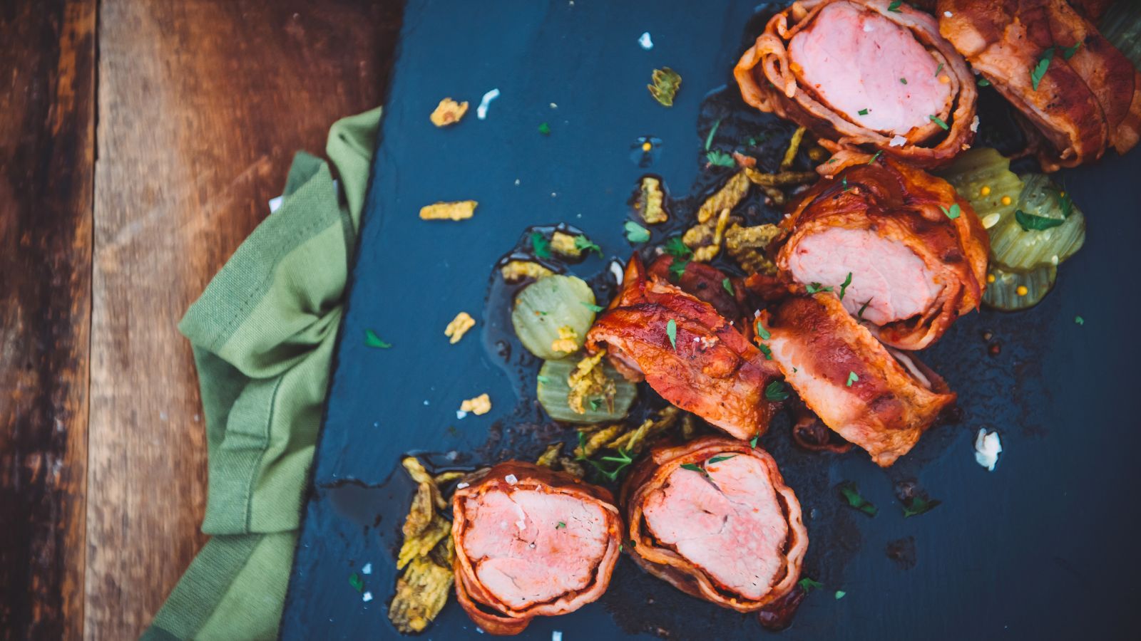 Slices of bacon-wrapped pork tenderloin are arranged on a dark slate board with roasted onions and herbs, next to a green cloth napkin on a wooden surface.