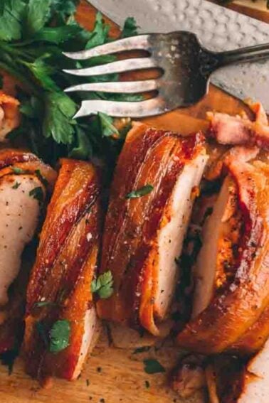 Sliced bacon-wrapped pork tenderloin on a wooden cutting board with herbs, a fork, and a knife.