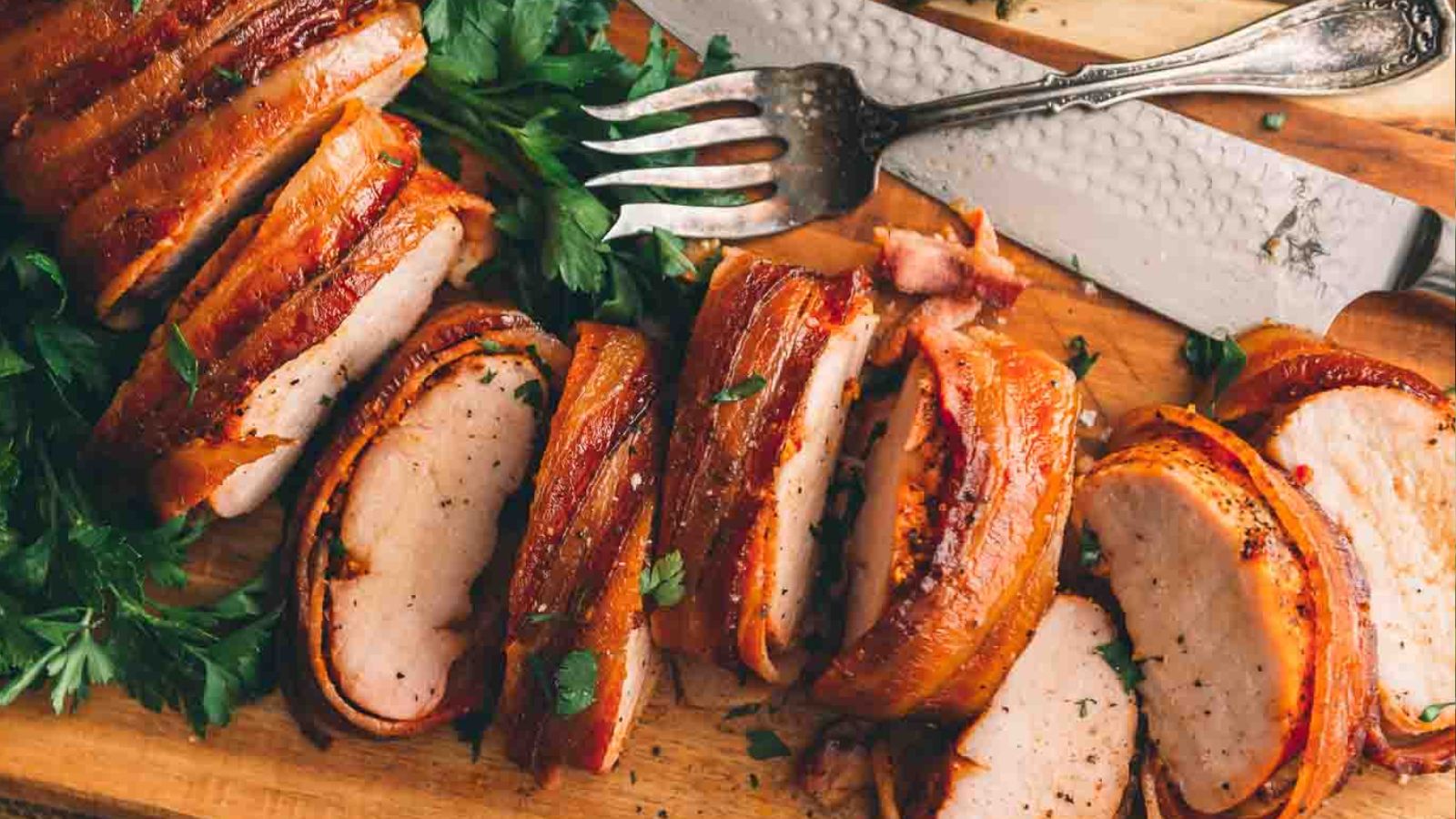 Sliced bacon-wrapped pork tenderloin on a wooden cutting board with herbs, a fork, and a knife.