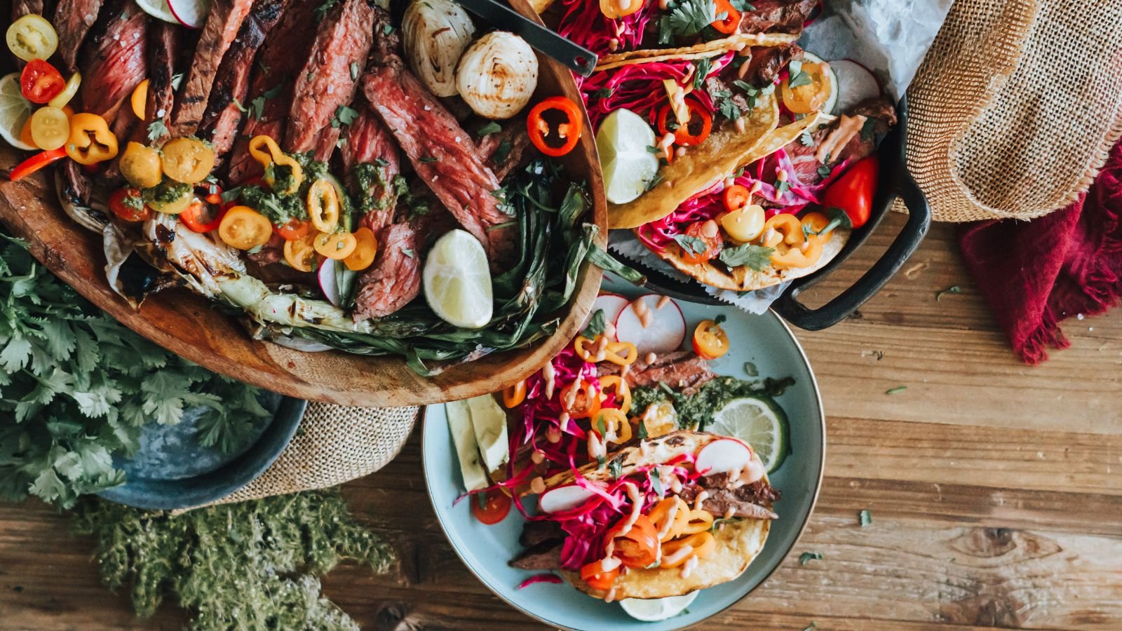 A wooden table with plates of sliced grilled steak, tacos topped with colorful vegetables, lime wedges, and fresh herbs.