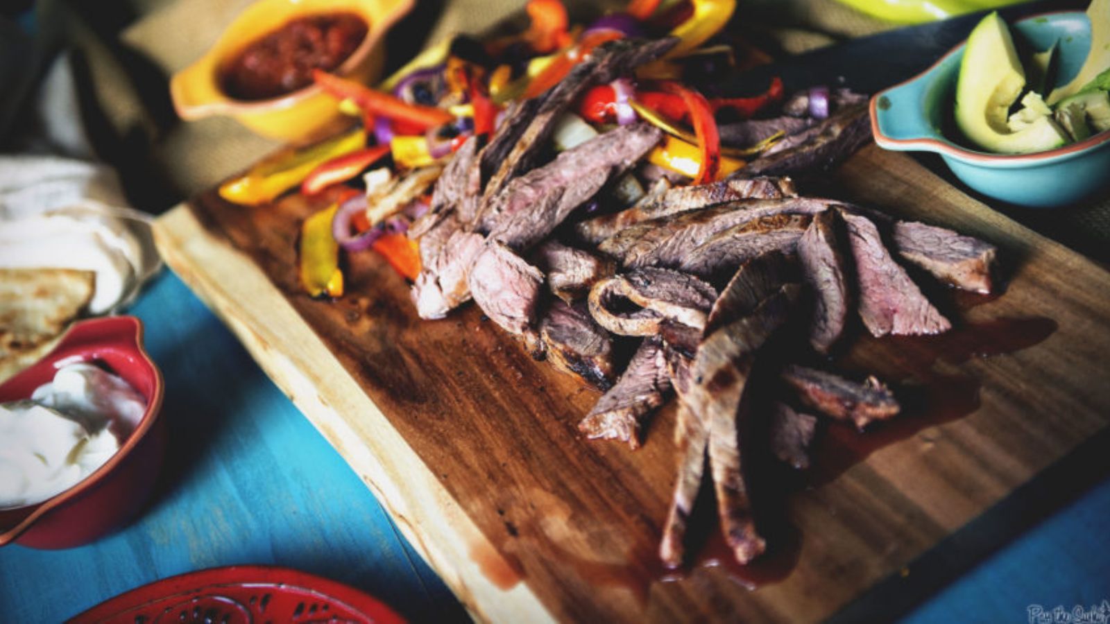 Sliced grilled steak on a wooden board with sautéed bell peppers and onions, surrounded by bowls of avocado, sour cream, and salsa.