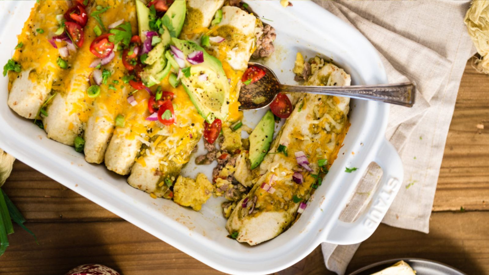 A white baking dish containing several enchiladas topped with melted cheese, avocado slices, diced tomatoes, red onion, and herbs, with a serving spoon inside.