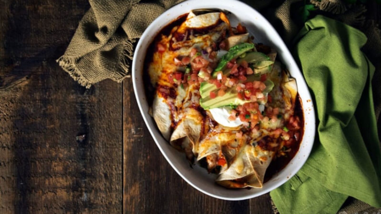 A white dish filled with baked enchiladas topped with melted cheese, avocado slices, pico de gallo, and sour cream, on a wooden table with green napkins.