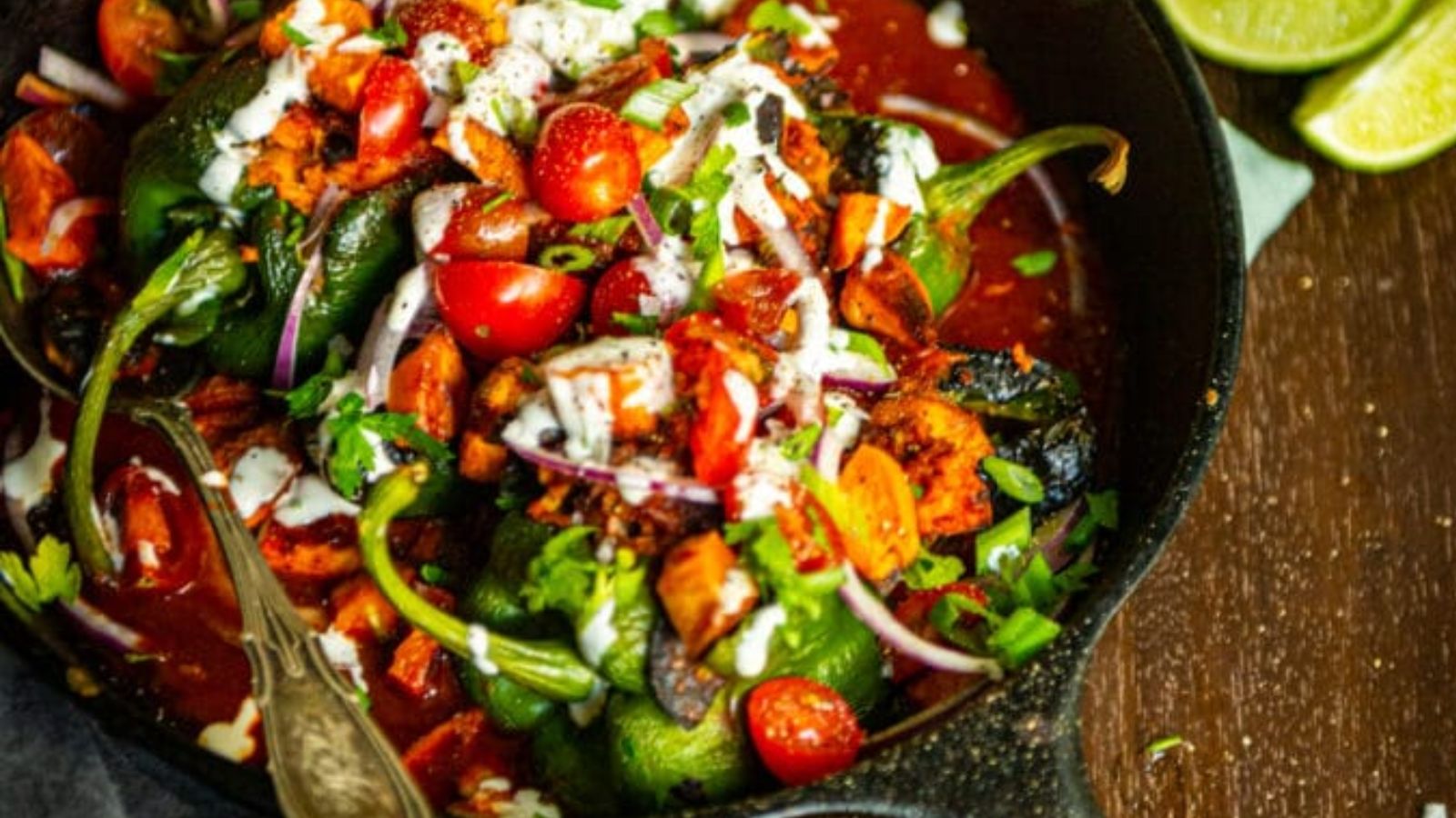 Stuffed poblano peppers topped with chopped tomatoes, onion, herbs, and a drizzle of white sauce in a cast iron skillet, with lime wedges on the side.